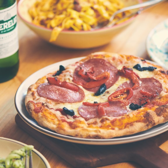 Porta Rocco pizza on a plate and table with pasta and green glass bottle in the background